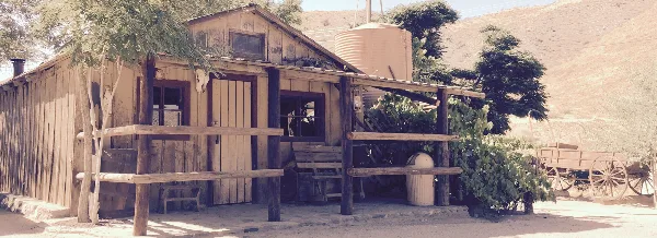 A rustic wooden ranch building with a porch, situated in a dry, hilly landscape with a water tank and wagon in the background.