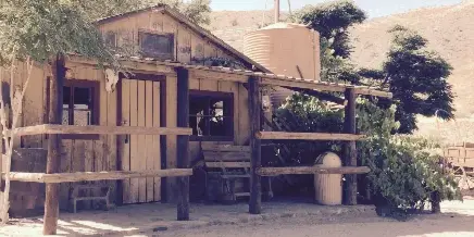 A rustic wooden ranch building with a porch, situated in a dry, hilly landscape with a water tank and wagon in the background.
