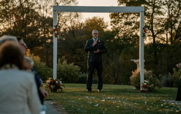 A wedding officiant in a suit stands at a white outdoor archway, speaking into a microphone to a seated audience during a ceremony.