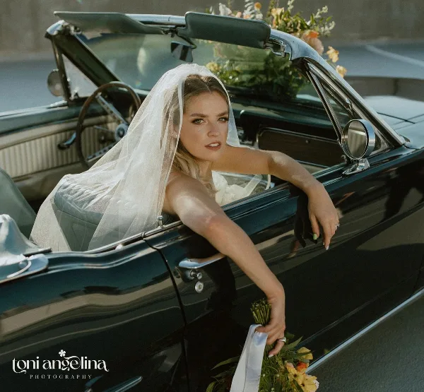 A bride with styled hair and makeup leaning out of a vintage convertible car.