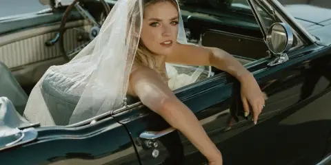 A bride with styled hair and makeup leaning out of a vintage convertible car.