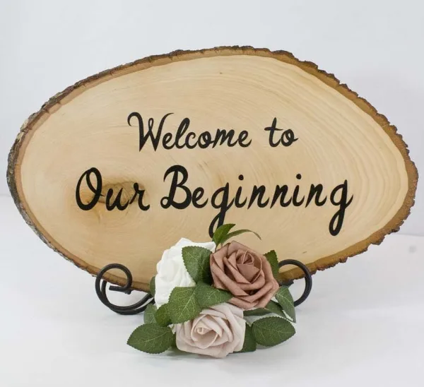 A rustic wooden slice sign reading 'Welcome to Our Beginning' decorated with faux roses and greenery.