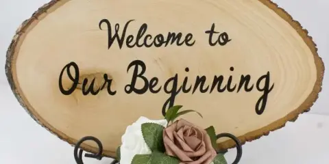 A rustic wooden slice sign reading 'Welcome to Our Beginning' decorated with faux roses and greenery.