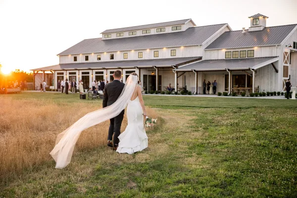 A bride and groom walking across a grassy field toward a white barn wedding venue at sunset.