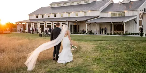 A bride and groom walking across a grassy field toward a white barn wedding venue at sunset.