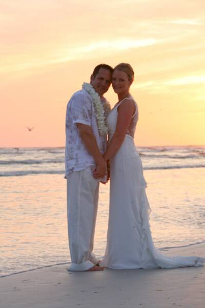 A bride and groom standing on a sandy beach at sunset, wearing white wedding attire.