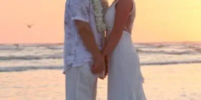 A bride and groom standing on a sandy beach at sunset, wearing white wedding attire.