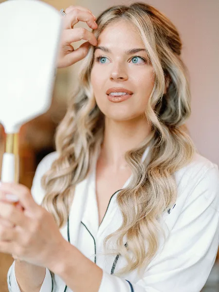 A bride with styled blonde hair and makeup looking into a hand mirror.