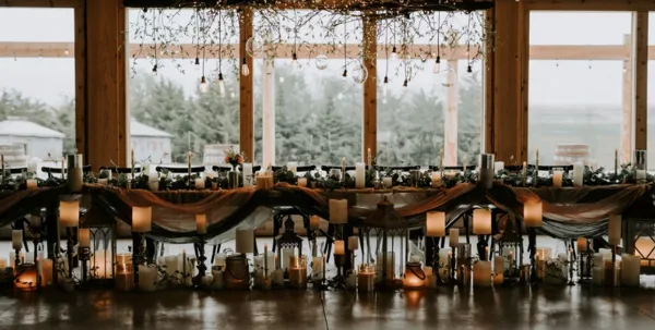 Rustic wedding reception table with candles and greenery inside a wood-framed event venue with large windows overlooking a countryside landscape.