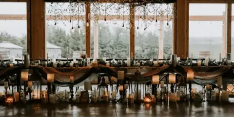 Rustic wedding reception table with candles and greenery inside a wood-framed event venue with large windows overlooking a countryside landscape.