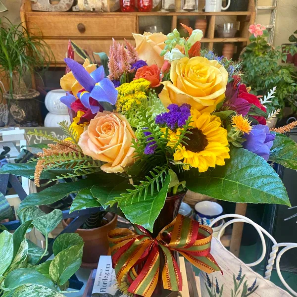 A colorful floral arrangement featuring yellow roses, a sunflower, and purple iris in a vase with a striped ribbon.