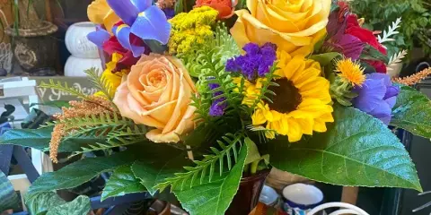A colorful floral arrangement featuring yellow roses, a sunflower, and purple iris in a vase with a striped ribbon.