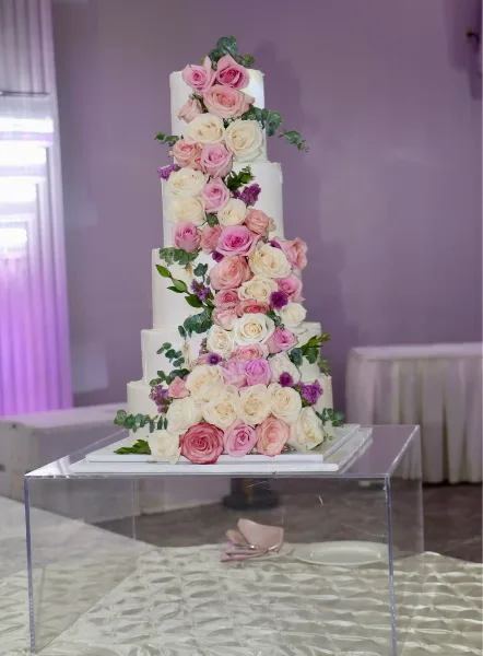 A tall, white multi-tiered wedding cake decorated with cascading pink and white roses and greenery, displayed on a clear acrylic stand.
