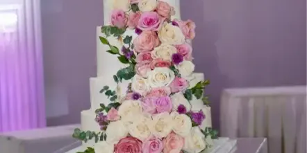 A tall, white multi-tiered wedding cake decorated with cascading pink and white roses and greenery, displayed on a clear acrylic stand.