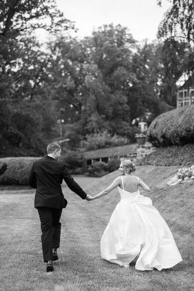 A black and white photograph of a bride and groom holding hands and walking across a grassy lawn in a garden setting.