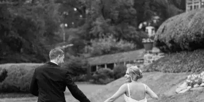 A black and white photograph of a bride and groom holding hands and walking across a grassy lawn in a garden setting.