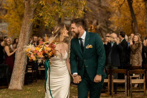 A bride and groom holding hands and smiling at each other during an outdoor wedding ceremony with guests in the background.