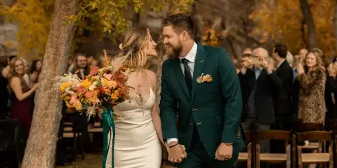 A bride and groom holding hands and smiling at each other during an outdoor wedding ceremony with guests in the background.