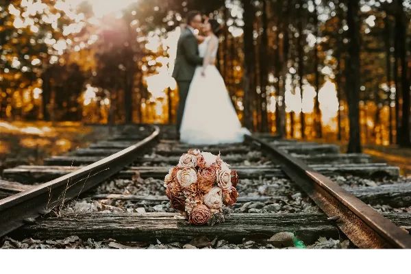A bride and groom standing on a wooded train track with a floral bouquet in the foreground.