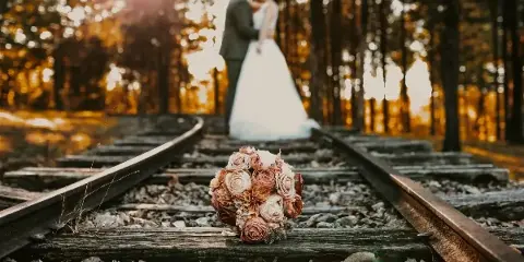 A bride and groom standing on a wooded train track with a floral bouquet in the foreground.