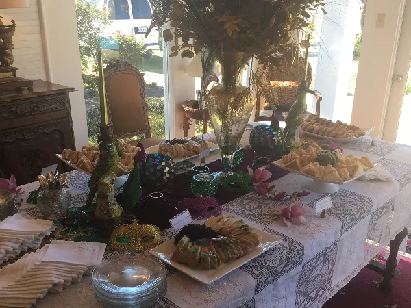 A catering table set with various food items including sandwiches, pastries, and a plated dish, decorated with floral arrangements and Mardi-themed ornaments.
