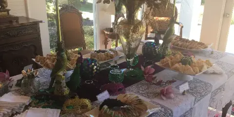 A catering table set with various food items including sandwiches, pastries, and a plated dish, decorated with floral arrangements and Mardi-themed ornaments.