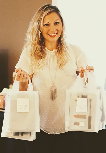 A woman holding two white tote bags labeled 'Janelle Bridal & Beauty' containing makeup products.