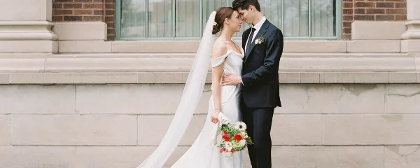 A bride and groom standing together in wedding attire in front of a stone building.