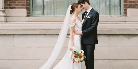 A bride and groom standing together in wedding attire in front of a stone building.
