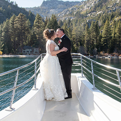 A bride and groom sharing a kiss on the deck of a boat with a mountainous lake landscape in the background.