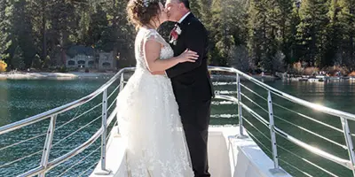 A bride and groom sharing a kiss on the deck of a boat with a mountainous lake landscape in the background.