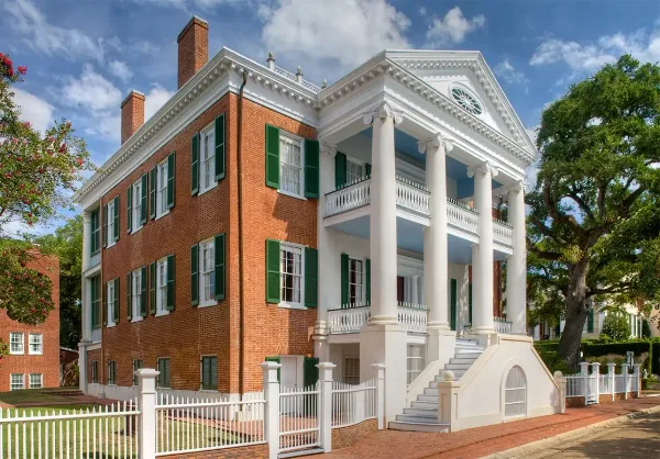 Exterior view of a historic red brick and white columned mansion with green shutters and a white picket fence.