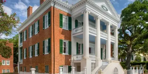 Exterior view of a historic red brick and white columned mansion with green shutters and a white picket fence.