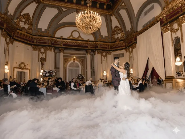 A bride and groom dancing on a fog-covered dance floor at an ornate wedding reception venue with seated guests.