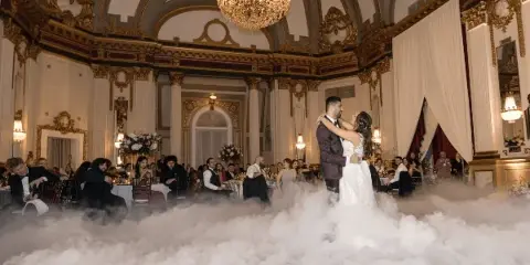 A bride and groom dancing on a fog-covered dance floor at an ornate wedding reception venue with seated guests.