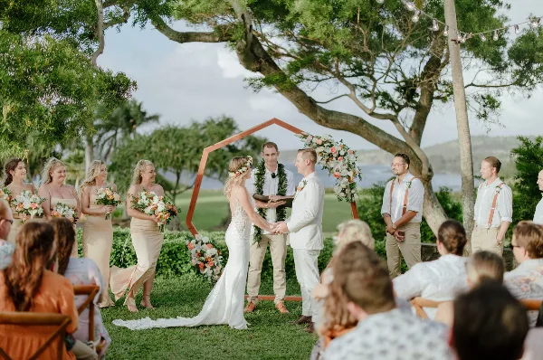 A wedding ceremony taking place outdoors under a large tree with a view of the ocean, featuring a bride and groom at an altar with a wedding party and guests.