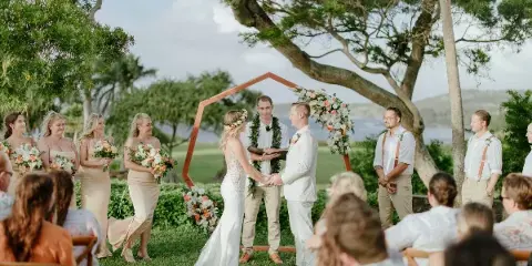 A wedding ceremony taking place outdoors under a large tree with a view of the ocean, featuring a bride and groom at an altar with a wedding party and guests.