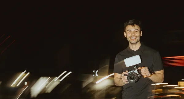 A smiling man holding a professional video camera with a microphone attachment against a dark background with light trails.
