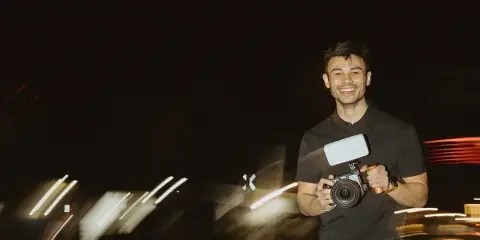 A smiling man holding a professional video camera with a microphone attachment against a dark background with light trails.