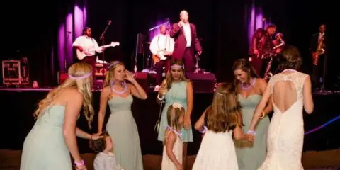 A live band performing on stage at a wedding reception with a bride and guests dancing in the foreground.