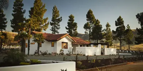 Exterior view of the white Spanish-style La Lomita Ranch estate with red tile roof, surrounded by palm trees and rolling hills.