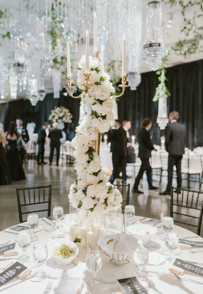 Luxury wedding reception table with white floral centerpiece, candles, and chandeliers in a ballroom.