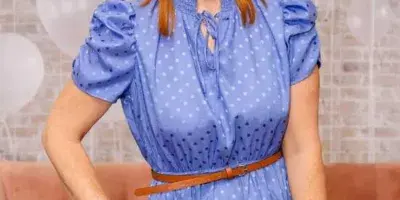 Portrait of a smiling woman with red hair wearing a blue polka-dot dress and a pearl headband, standing in front of a brick wall and white balloons.
