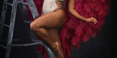 A woman in a white outfit and red feathered wings posing on a ladder in a studio setting.