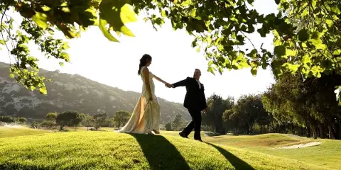 Bride and groom holding hands on a grassy hillside with trees and mountains in the background