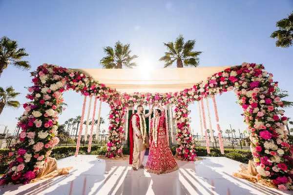 A couple in traditional Indian wedding attire standing under a large, custom-built floral mandap structure outdoors.