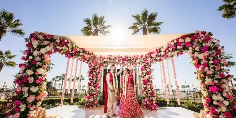 A couple in traditional Indian wedding attire standing under a large, custom-built floral mandap structure outdoors.