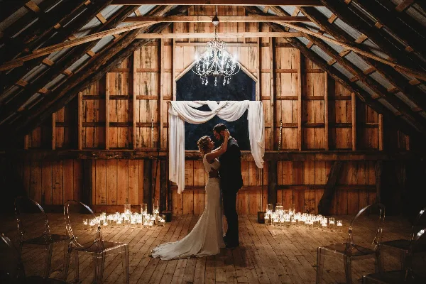 A bride and groom dancing in a rustic wooden barn with a chandelier and candles.