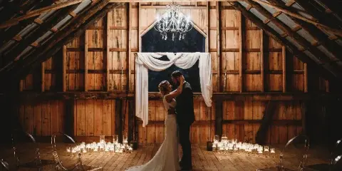 A bride and groom dancing in a rustic wooden barn with a chandelier and candles.