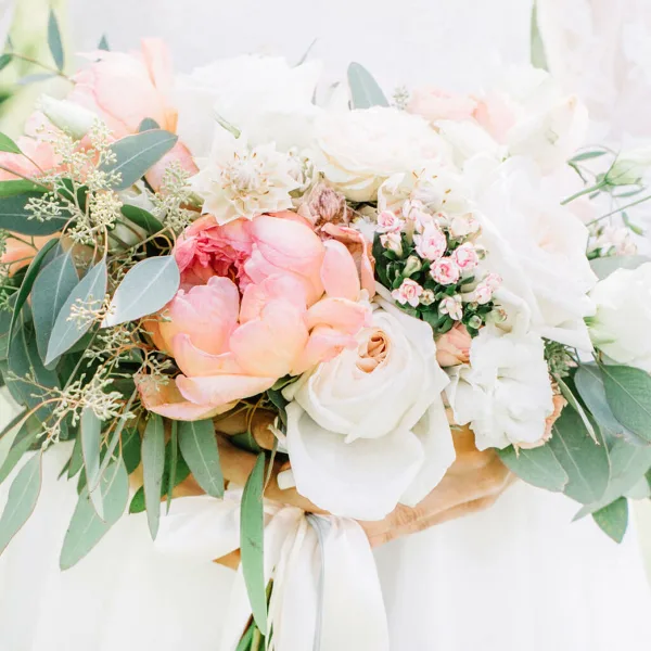 A close-up photograph of a floral bouquet featuring white and peach roses, ranunculus, and eucalyptus leaves.